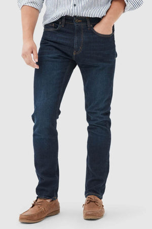 Wearing Rodd & Gunn Sutton Straight Italian Denim Jeans, a striped shirt with rolled-up sleeves, and brown boat shoes, a person stands with one hand in their pocket against a plain light background.