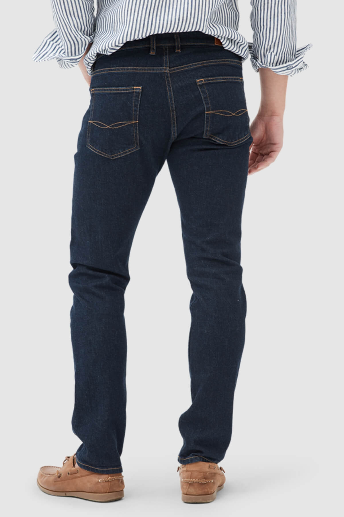 A person wearing Rodd & Gunn Sutton Straight Italian Denim Jeans, a blue-and-white striped shirt with rolled-up sleeves, and light brown moccasin shoes, seen from the back against a plain light background.