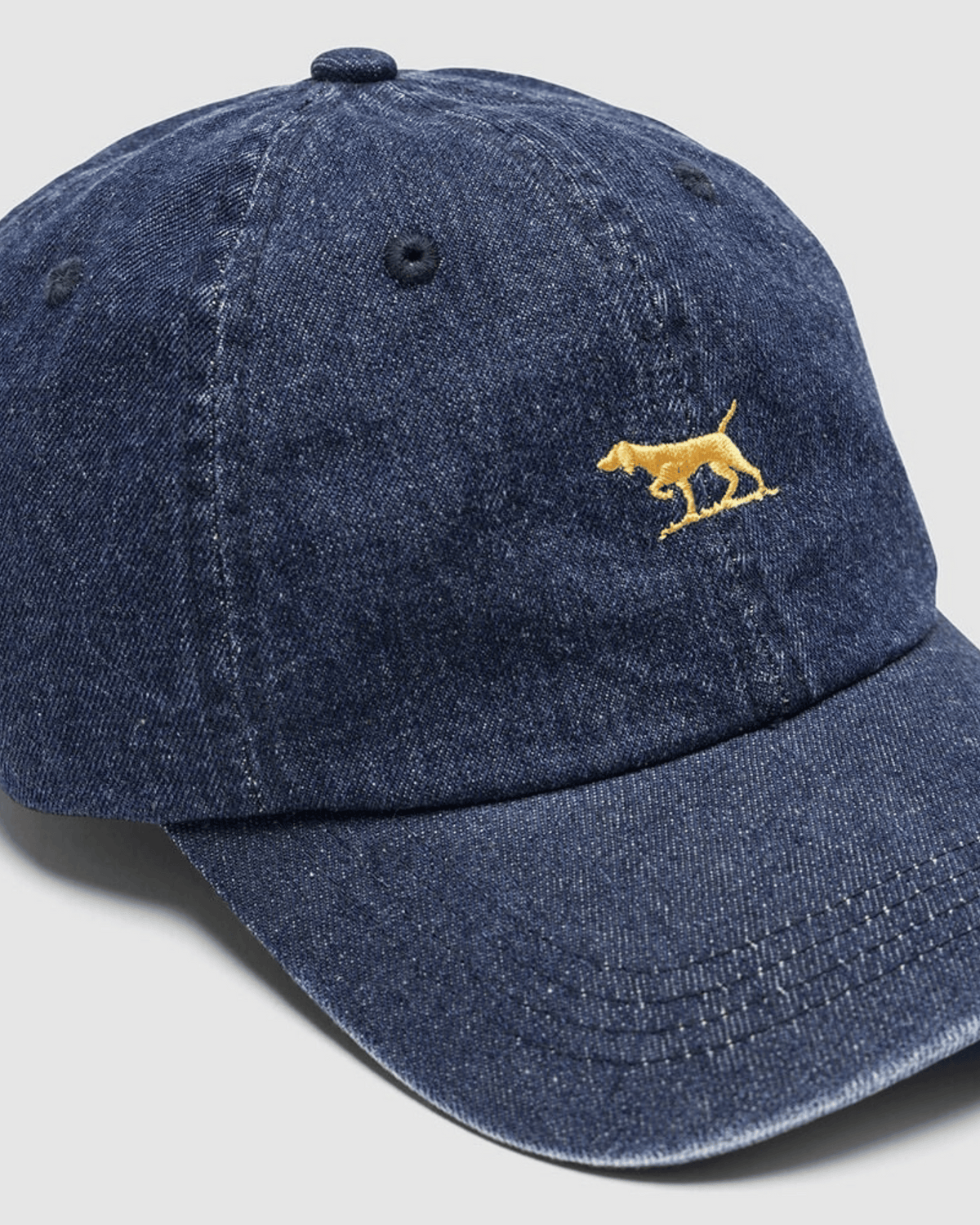 The Rodd & Gunn Gunn Cap is a blue denim hat made from durable cotton twill, with a small yellow embroidered dog on the front, shown on a light gray background.
