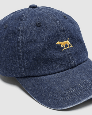 The Rodd & Gunn Gunn Cap is a blue denim hat made from durable cotton twill, with a small yellow embroidered dog on the front, shown on a light gray background.