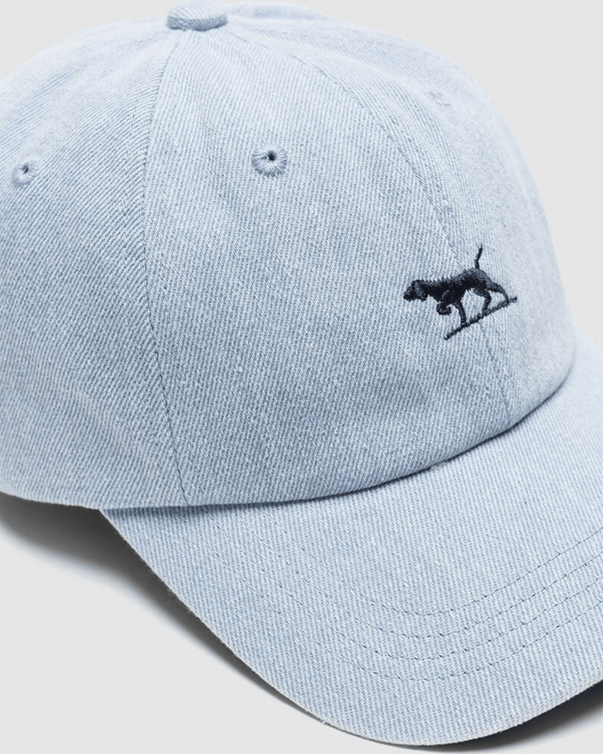 The Rodd & Gunn Gunn Cap is made from heavy duty cotton twill in light blue denim and features a small black embroidered dog on the front.