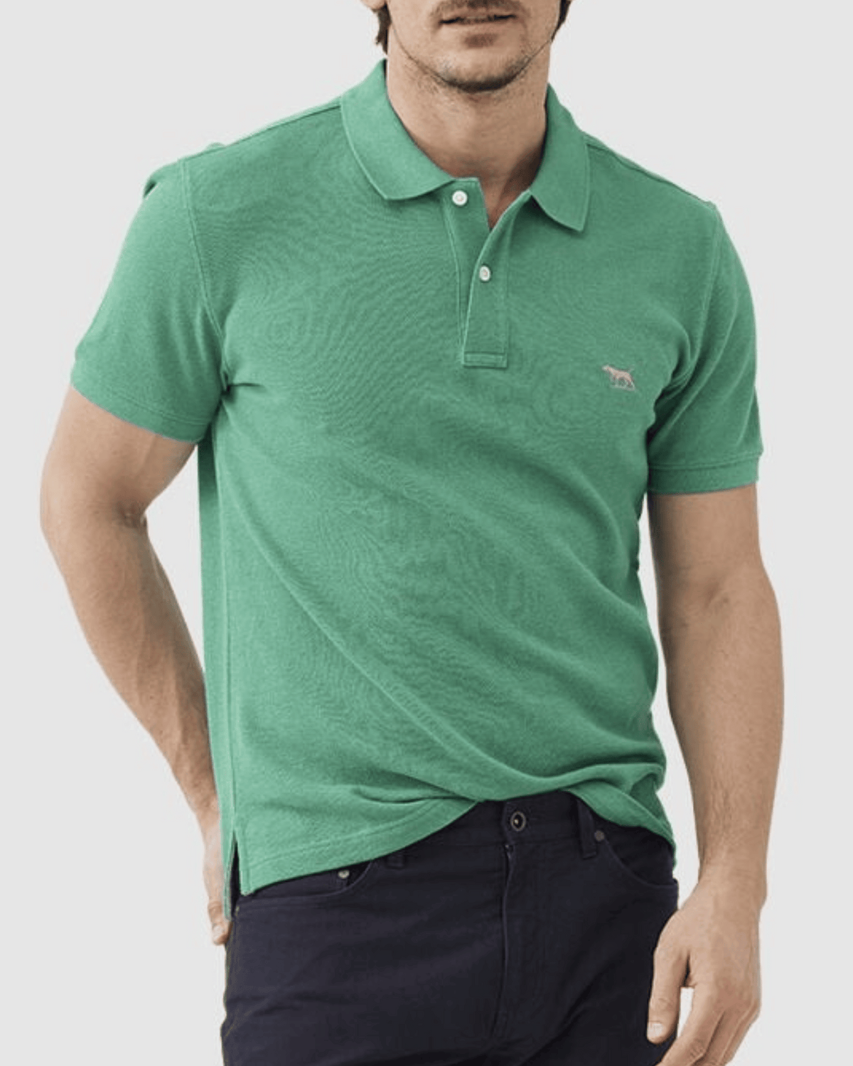 A person in a Rodd & Gunn The Gunn Polo, made from Australian cotton with an embroidered chest logo, and dark pants stands with one hand in their pocket against a plain background.