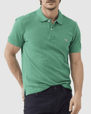 A person in a Rodd & Gunn The Gunn Polo, made from Australian cotton with an embroidered chest logo, and dark pants stands with one hand in their pocket against a plain background.