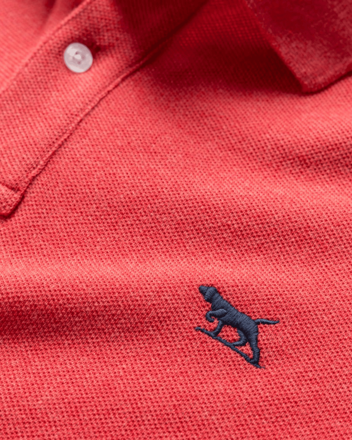Close-up of the Rodd & Gunn The Gunn Polo, a modern fit red textured shirt made from soft Australian cotton, with a white button and a small navy blue embroidered dog logo on the chest.
