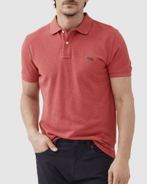 A man wearing the Rodd & Gunn The Gunn Polo in modern fit, short sleeves, and red Australian cotton stands with one hand in his pocket, paired with dark pants against a plain light background.