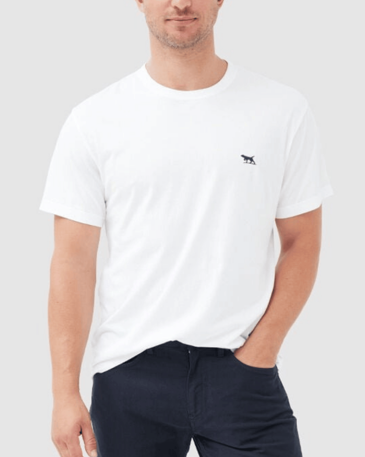 A person wears the Rodd & Gunn The Gunn T-Shirt, a plain white short-sleeve made from Australian Cotton with a small chest logo. Paired with sports-fit dark pants, they stand against a light gray background, one hand in their pocket, embodying effortless style.