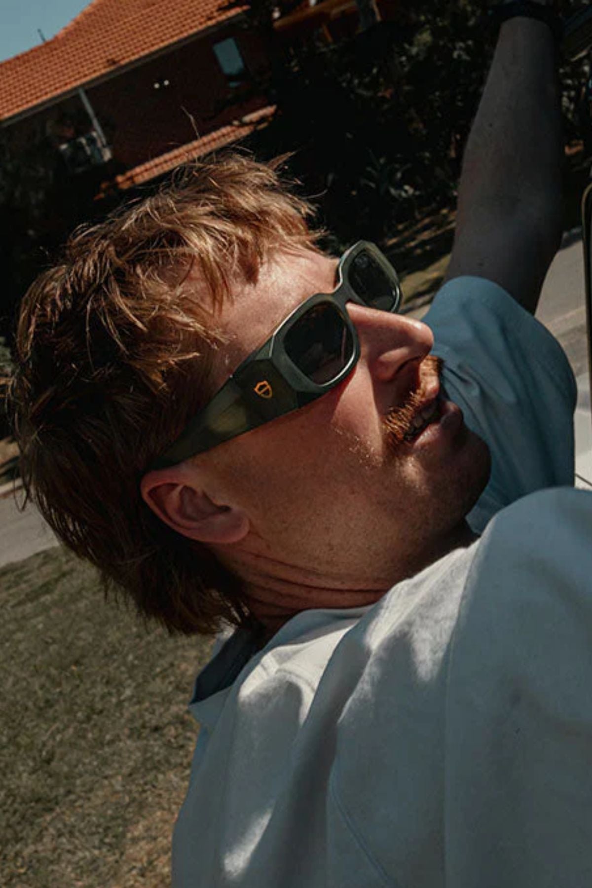 A man with light brown hair and a mustache wears SafeStyle Auras Matte Moss Green Frame Grey Polarised Lens safety glasses and a white shirt, looking upward outdoors on a sunny day with a red-tiled roof house in the background.