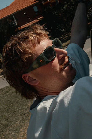 A man with light brown hair and a mustache wears SafeStyle Auras Matte Moss Green Frame Grey Polarised Lens safety glasses and a white shirt, looking upward outdoors on a sunny day with a red-tiled roof house in the background.
