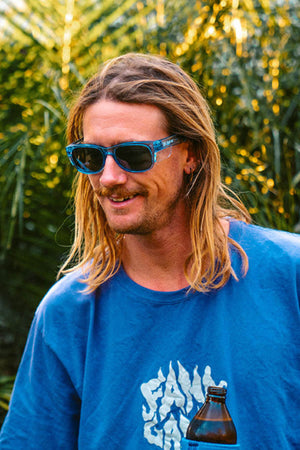 A person with long blonde hair and a mustache enjoys the sun in a blue T-shirt, wearing SafeStyle Classics Blue Frame Polarised Lens sunglasses. They smile while holding a bottle, basking in sunlight filtered through greenery, appreciating the UV protection benefits.