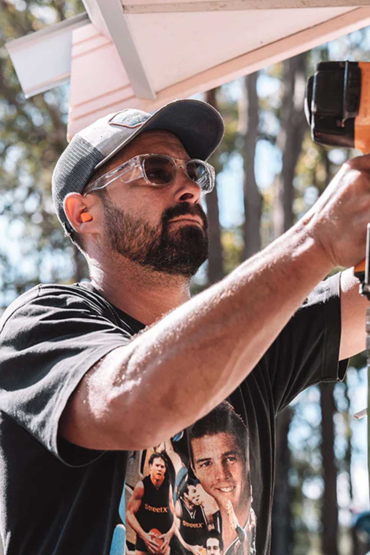 A person wearing SafeStyle Classics Clear Frame Hybrids with photochromic lenses and a cap uses a power drill outdoors while working on a structure. Trees are in the background, and their black T-shirt sports a graphic design.