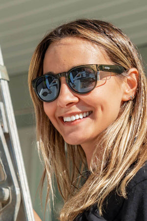A woman with long, light brown hair smiles while wearing SafeStyle Cruisers sunglasses with green frames and polarized lenses. She stands by a metal object, possibly a ladder, against a light background. Her sunglasses reflect the surroundings and provide UV protection.