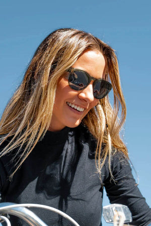 A smiling woman with long blonde hair wears SafeStyle Cruisers, green frame polarized lens sunglasses, featuring UV protection and polycarbonate frames, perfectly complementing her black top against the clear blue sky.