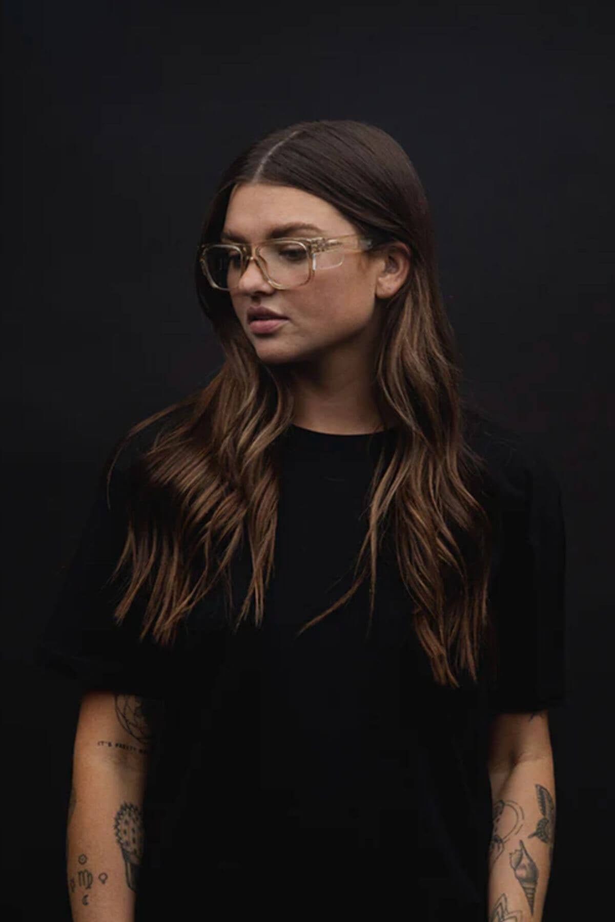 Against a dark backdrop, a person with long, wavy hair wears a black t-shirt and SafeStyle Fusions Champagne Frame Glasses. Showcasing arm tattoos, their neutral expression is visible through the lenses designed for blue light protection.