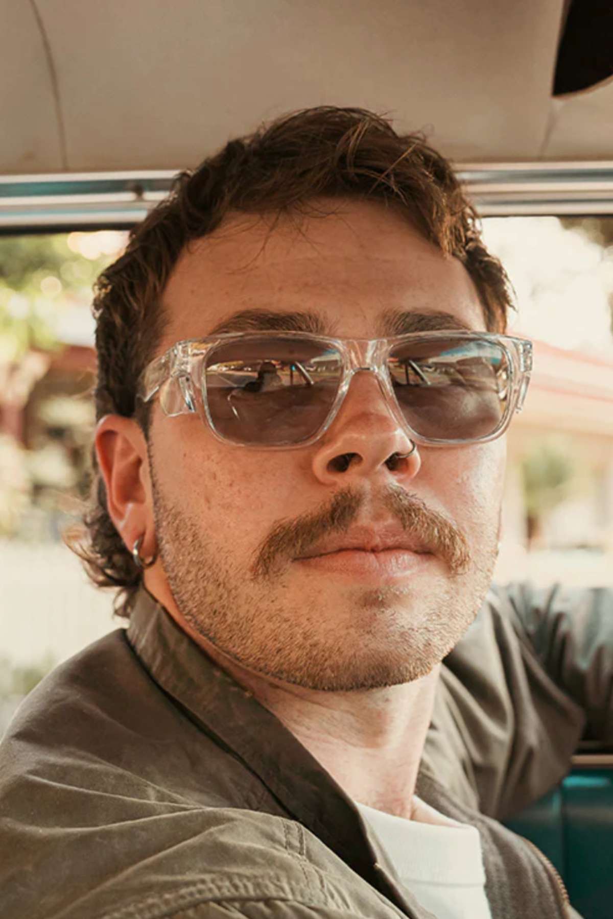 A person with short curly hair and a mustache sits in a car, wearing SafeStyle Fusions Clear Frame Hybrids featuring UV protection and shatterproof polycarbonate lenses. An earring glints as they look away from the camera against a softly blurred background.