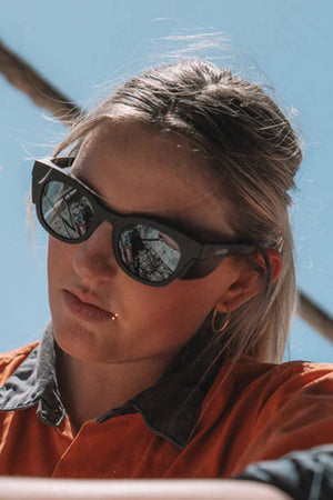 A blonde-haired person wears SafeStyle Harps Matte Black Frame Polarised Lens sunglasses with UV protection and an orange collared shirt. A blue sky, beams, and a reflection of a metal structure appear in the background.