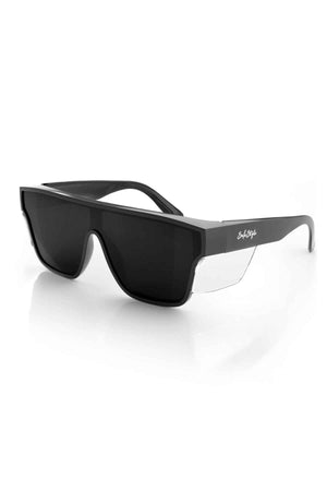 SafeStyle Primes Black Frame Polarised Lens sunglasses are a stylish pair of black, flat-top shades with dark lenses. They feature thick black frames and clear transparent sides near the hinges, made from shatterproof polycarbonate. The brand's signature adorns the temple arms, and they are displayed on a white reflective surface.