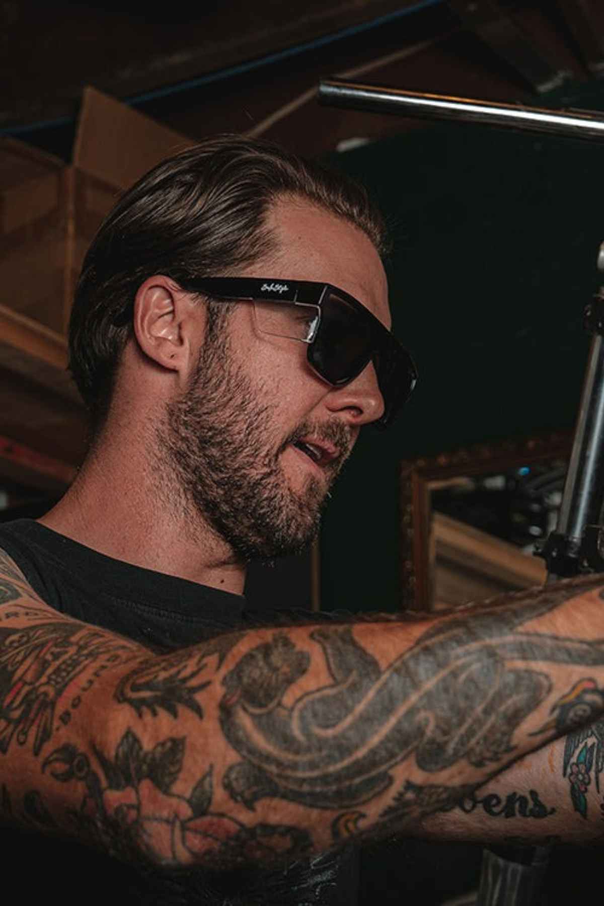 A man wearing SafeStyle Primes with black polycarbonate frames and polarised UV400 lenses, sporting tattoos on his arms, is working on a piece of equipment in a dimly lit room. He has a beard and short hair and appears focused on the task at hand.