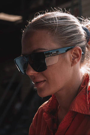 A blonde-haired woman in a ponytail wears SafeStyle Primes Black Frame Tinted Lens sunglasses and a red shirt, standing in a dimly lit workshop or industrial setting.