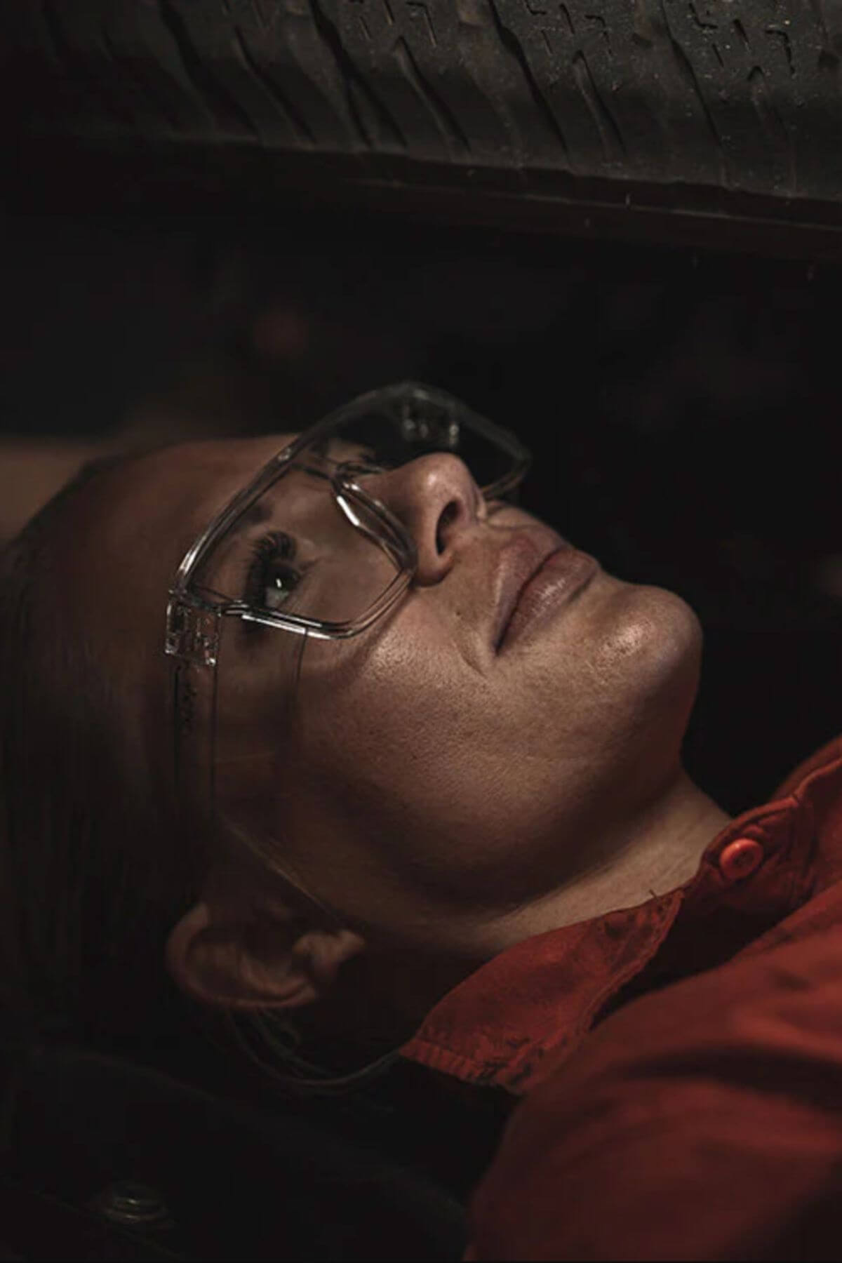 Wearing SafeStyle Primes Clear Frame Clear Lens safety glasses and a red shirt, a person lies beneath a vehicle tire, looking up as if inspecting or repairing something under the vehicle.