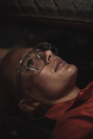 Wearing SafeStyle Primes Clear Frame Clear Lens safety glasses and a red shirt, a person lies beneath a vehicle tire, looking up as if inspecting or repairing something under the vehicle.