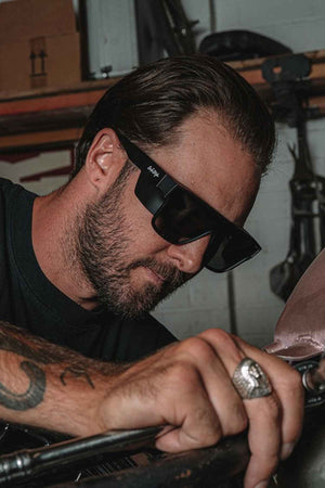 In a workshop, a bearded man wearing SafeStyle Primes Matte Black Frame Polarised Lens sunglasses focuses on his mechanical project. A tattooed arm and an "S" ring add to his look as he works with precision and dedication.