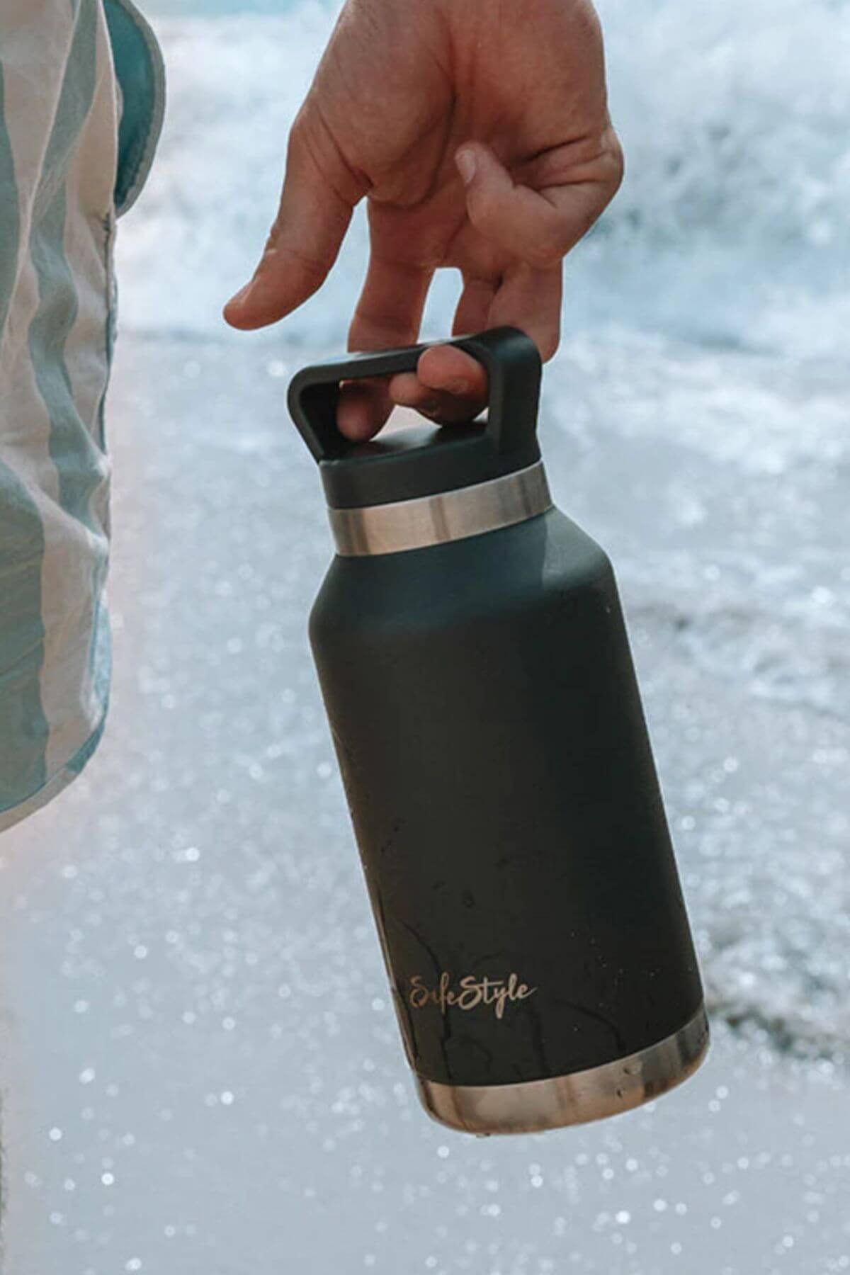 A person holds a leak-proof black and silver Safestyle Big Juicy 1L Water Bottle, featuring double-wall vacuum insulation above ocean waves. They wear a blue and white striped garment against the backdrop of a sandy beach with shimmering water.