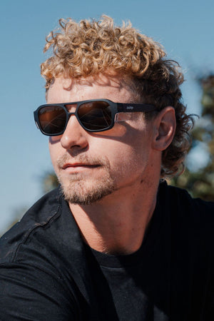 A man with curly blond hair and a short beard wears Safestyle Skylines sunglasses featuring a chocolate frame and brown polarised lenses, paired with a black shirt, looking to the side outdoors under a sunny blue sky.