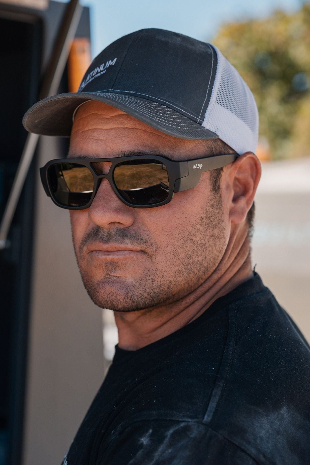 A man wearing Safestyle Skylines Matte Black Frame sunglasses with grey polarised lenses, a black T-shirt, and a gray and white trucker hat looks at the camera outdoors on a sunny day.