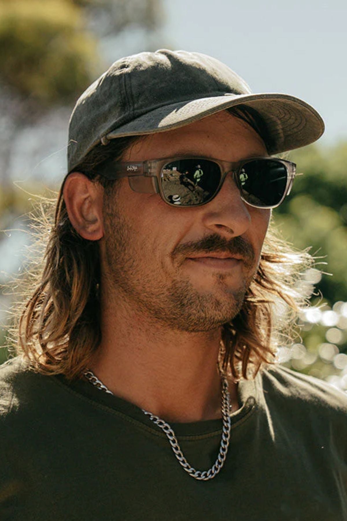 A man with long hair, a green cap, and a dark green T-shirt stands outdoors on a sunny day wearing Safestyle Classics Matte Graphite Frame Polarised Lens sunglasses, a silver chain necklace; trees and sky blur in the background.