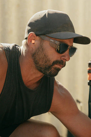 A bearded man in a black cap, sleeveless shirt, and Safestyle Fusions Matte Graphite Frame Polarised sunglasses is working outdoors in bright sunlight.
