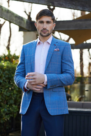 A man with short dark hair and a beard stands outdoors in the Savile Row Abram Wool Blazer, paired with a light pink dress shirt and dark blue trousers. He looks at the camera, greenery and wooden beams behind him.
