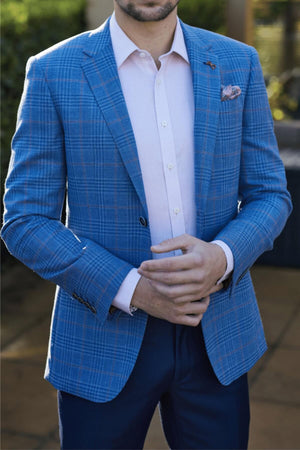 A man stands outdoors, adjusting his cuff while wearing the Savile Row Abram Wool Blazer—a blue plaid slim-fit jacket over a pale pink dress shirt and finished with a pocket square. His face is partially out of frame.