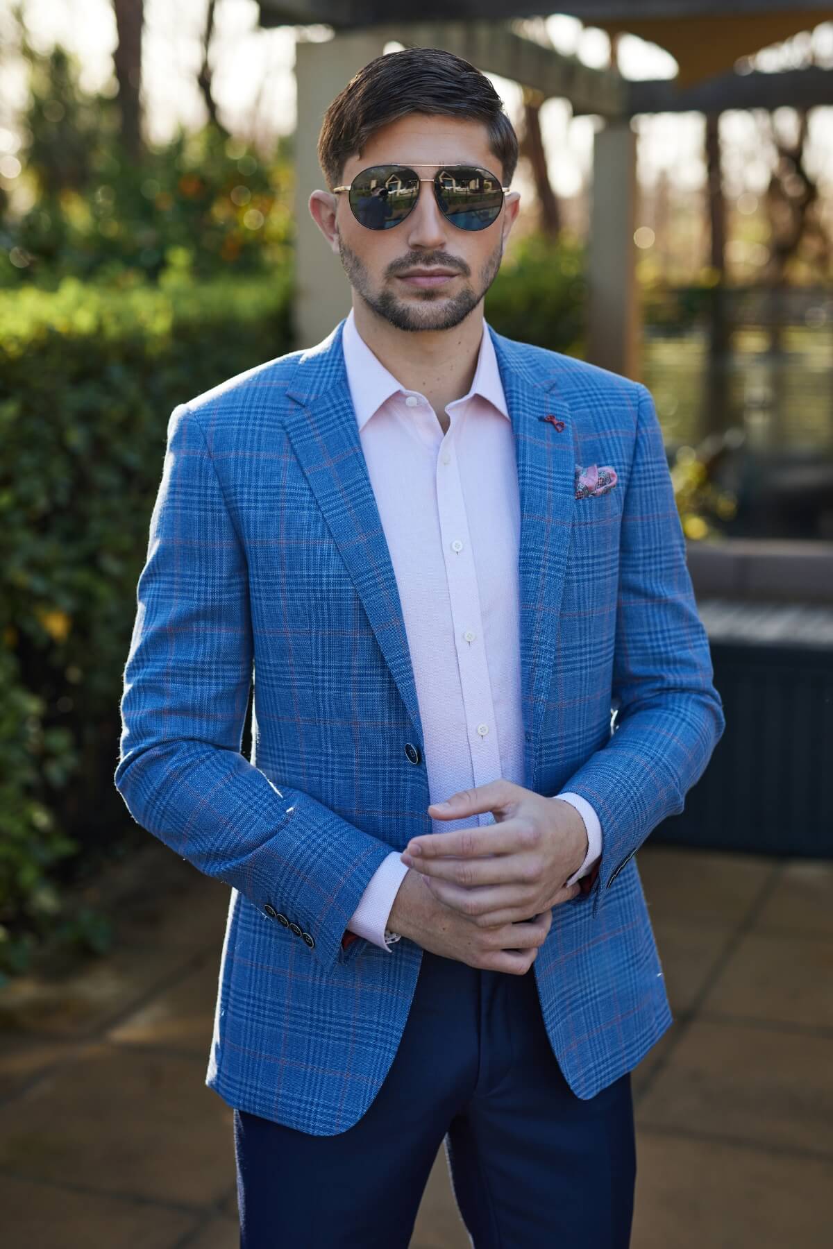 A man with short dark hair and a beard wears aviator sunglasses, a light pink dress shirt, dark trousers, and the Savile Row Abram Wool Blazer. He stands outdoors on a sunny day with greenery in the background.