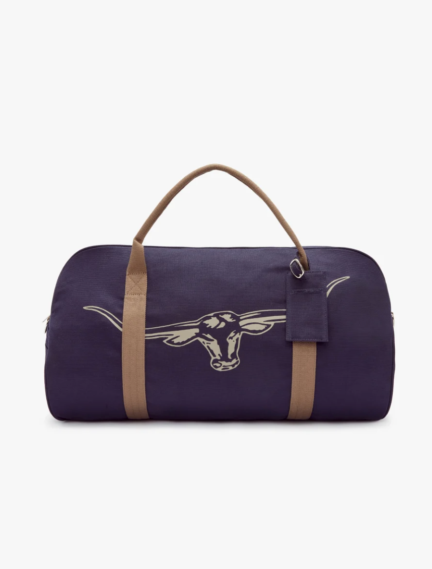 The RM Williams Nanga Canvas Bag is a dark blue duffel with a white longhorn emblem on the front. Made from durable cotton, it includes an exterior pocket, beige handles, a matching detachable shoulder strap, and an attached luggage tag on a plain white background.