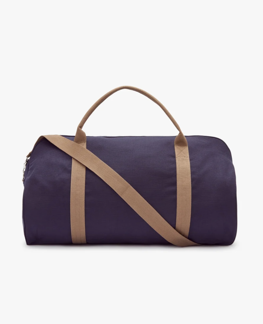 The RM Williams Nanga Canvas Bag, in navy blue with durable cotton fabric and tan straps, is shown against a white background. It includes two short handles, an adjustable shoulder strap, and an exterior pocket for convenience.