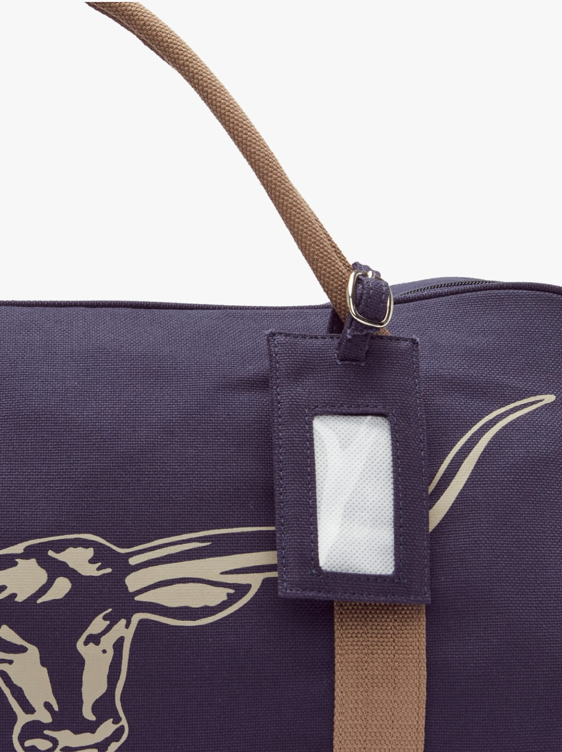 Close-up of the RM Williams Nanga Canvas Bag, a dark cotton bag with brown handles, an exterior pocket, a steer head design, plus a tag holder near the zipper.