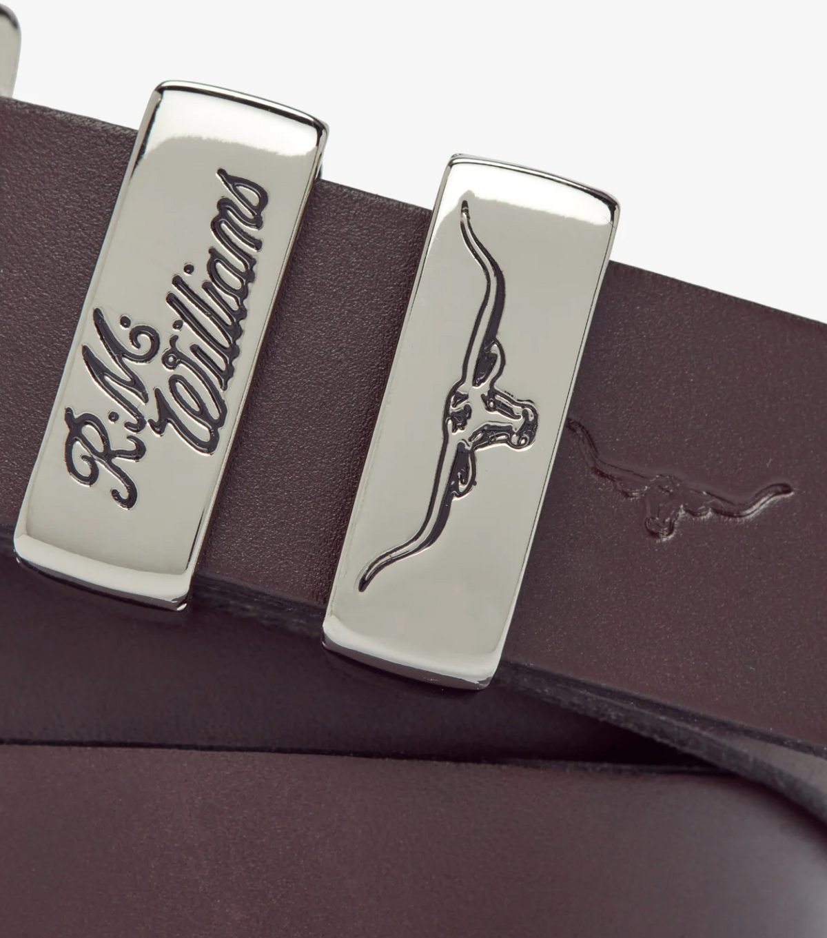Close-up of the RM Williams Slim Drover Belt in solid brown hide leather with two silver nickel buckles—one engraved "R.M. Williams," the other with a longhorn cattle logo. The logo is also embossed on the leather. Made in Australia.