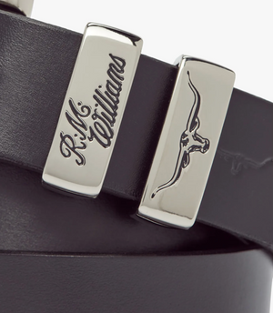 Close-up of the RM Williams Slim Drover Belt in black solid hide leather, with two silver accents: one engraved "R.M.Williams" in script, the other showing a longhorn bull head, complemented by a classic nickel buckle.