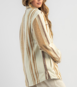 A person with long hair wears a Rip Curl Kindred Palms Jacket in cream and brown stripes, standing sideways to highlight the polyester shacket's back and side. The background is plain light gray.