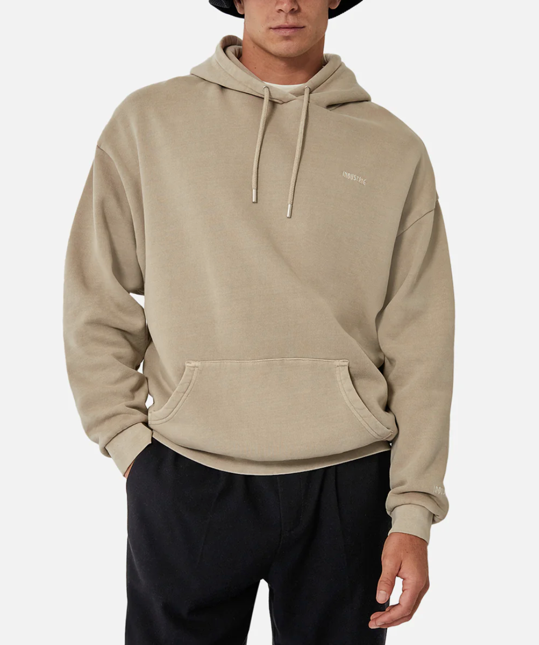 A person wearing the Industrie The Del Sur Hoodie in an oversized beige fit with a drawstring hood, subtle chest text, front pocket, and black pants stands against a plain white background, face partially visible.