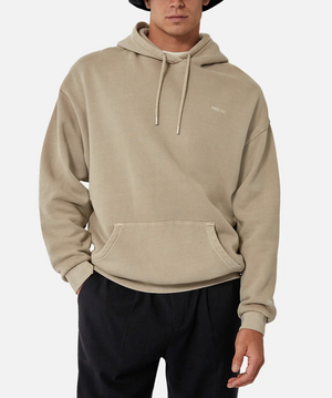 A person wearing the Industrie The Del Sur Hoodie in an oversized beige fit with a drawstring hood, subtle chest text, front pocket, and black pants stands against a plain white background, face partially visible.