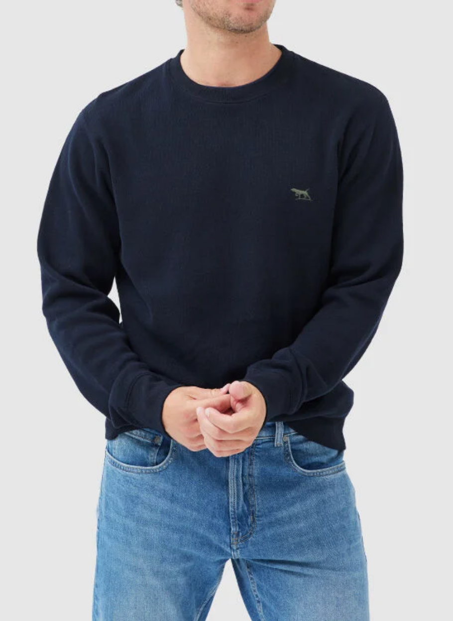 A person, head cropped, stands against a light background wearing the Rodd & Gunn Gunn Crew Neck Sweat in dark navy 100% cotton with a relaxed fit and small animal logo on the left chest, paired with light blue jeans.