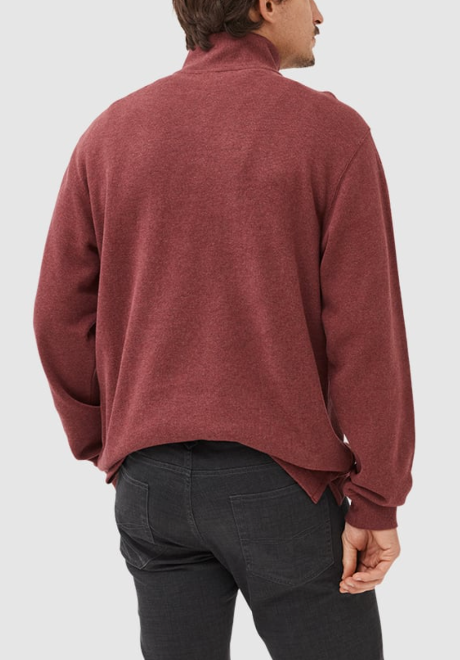 A man in a maroon Rodd & Gunn Alton Ave Sweat, made of 100% cotton and featuring a slightly high collar, stands with his back to the camera. He pairs it with black pants and the sweater hem is unevenly tucked. The background is plain gray.