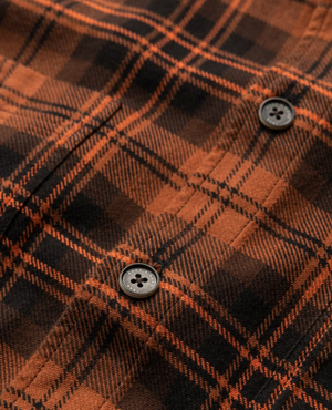 Close-up of the Rodd & Gunn Bryant Park Shirt, a stylish orange and black plaid flannel made from 100% cotton. Features a checkered pattern with diagonal lines, visible stitching, and black buttons.