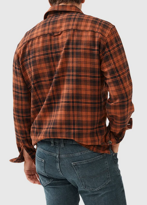 A person is seen from behind wearing the Rodd & Gunn Bryant Park Shirt, a stylish orange and black plaid flannel made of 100% cotton, with partially rolled-up sleeves, paired with blue jeans and one hand in their back pocket.