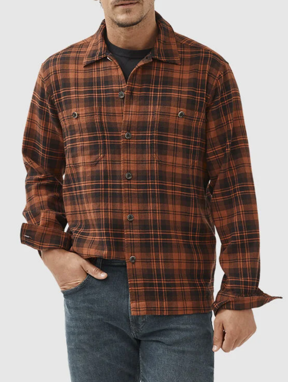 A man wearing the Rodd & Gunn Bryant Park Shirt—a stylish, 100% cotton brown and black plaid button-up with chest pockets—and blue jeans stands with one hand in his pocket against a plain white background.
