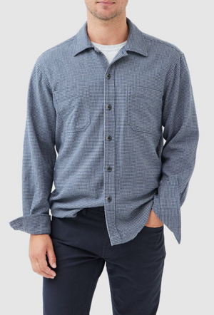 A person in a Rodd & Gunn Pentland Hills blue and black checkered long-sleeve shirt, made of 100% cotton, stands against a white background. The shirt has front buttons and two chest pockets, while their hand rests in their dark pants pocket.
