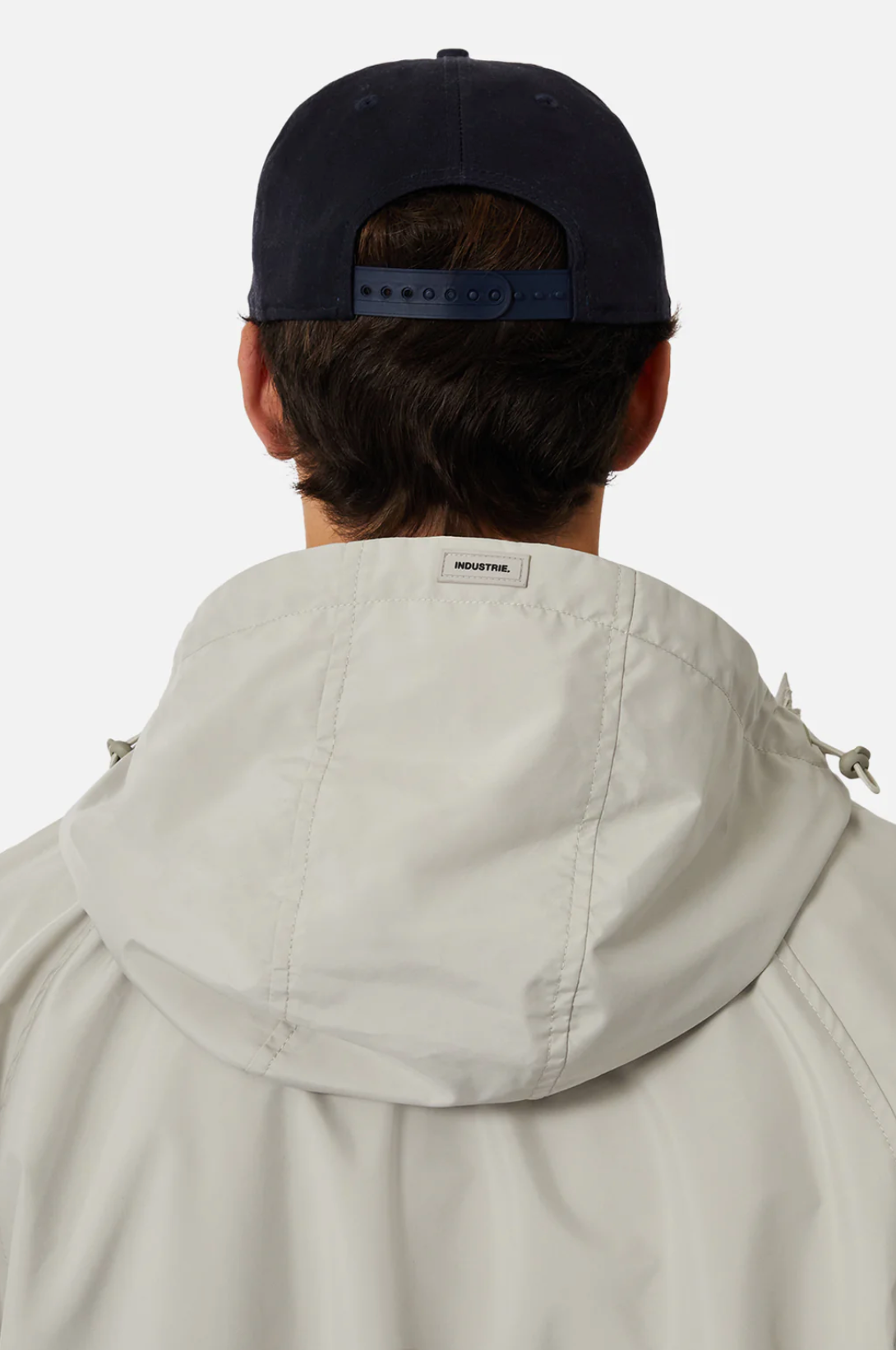A person is seen from behind wearing a dark cap and light Industrie The Rain Jacket with the logo on the hood, set against a plain white background that highlights their simple attire.