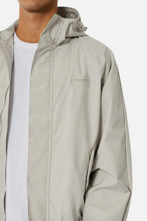 A person is shown from shoulders to waist in a light beige Industrie The Rain Jacket with the logo subtly visible on the chest over a white T-shirt, set against a plain white background.