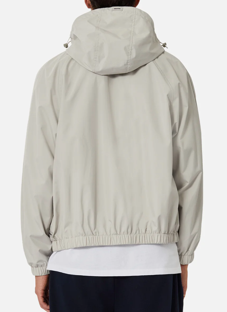 A person is seen from behind wearing Industrie The Rain Jacket in light gray, featuring a drawstring hood and elastic waistband. A white shirt underneath subtly contrasts with dark pants. The jacket is branded with the Industrie logo.